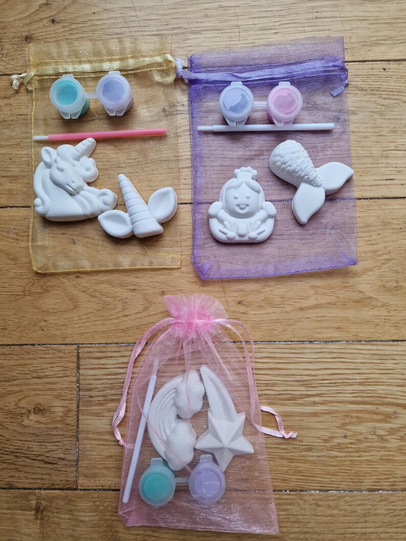 Puede incluir: Tres bolsas de organza llenas de pintura, pinceles y figuritas de cer&aacute;mica para pintar. Las bolsas son amarillas, moradas y rosas. Las figuritas incluyen un unicornio, una sirena, una princesa, un arco&iacute;ris y una estrella.