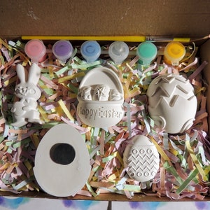 May include: A craft kit with five white ceramic Easter-themed figurines, including a bunny, a basket, and three eggs. The kit also includes six small bottles of paint and a paintbrush. The figurines are sitting on a bed of colorful shredded paper in a cardboard box.