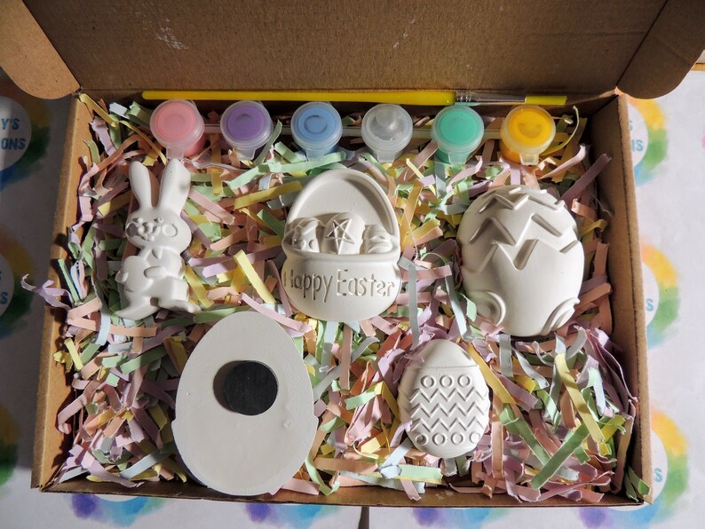 May include: A craft kit with five white ceramic Easter-themed figurines, including a bunny, a basket, and three eggs. The kit also includes six small bottles of paint and a paintbrush. The figurines are sitting on a bed of colorful shredded paper in a cardboard box.