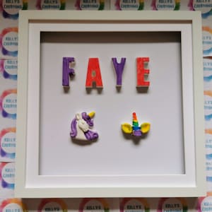 May include: A white picture frame with a white mat. The name "FAYE" is spelled out in 3D letters in purple, red, and purple. There are two unicorn decorations, one purple and one rainbow colored.