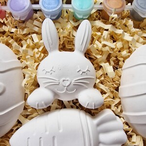 May include: A set of four white ceramic Easter-themed figurines, including a bunny, an egg, and two carrots, ready to be painted. The set comes with six small pots of paint in various colors.
