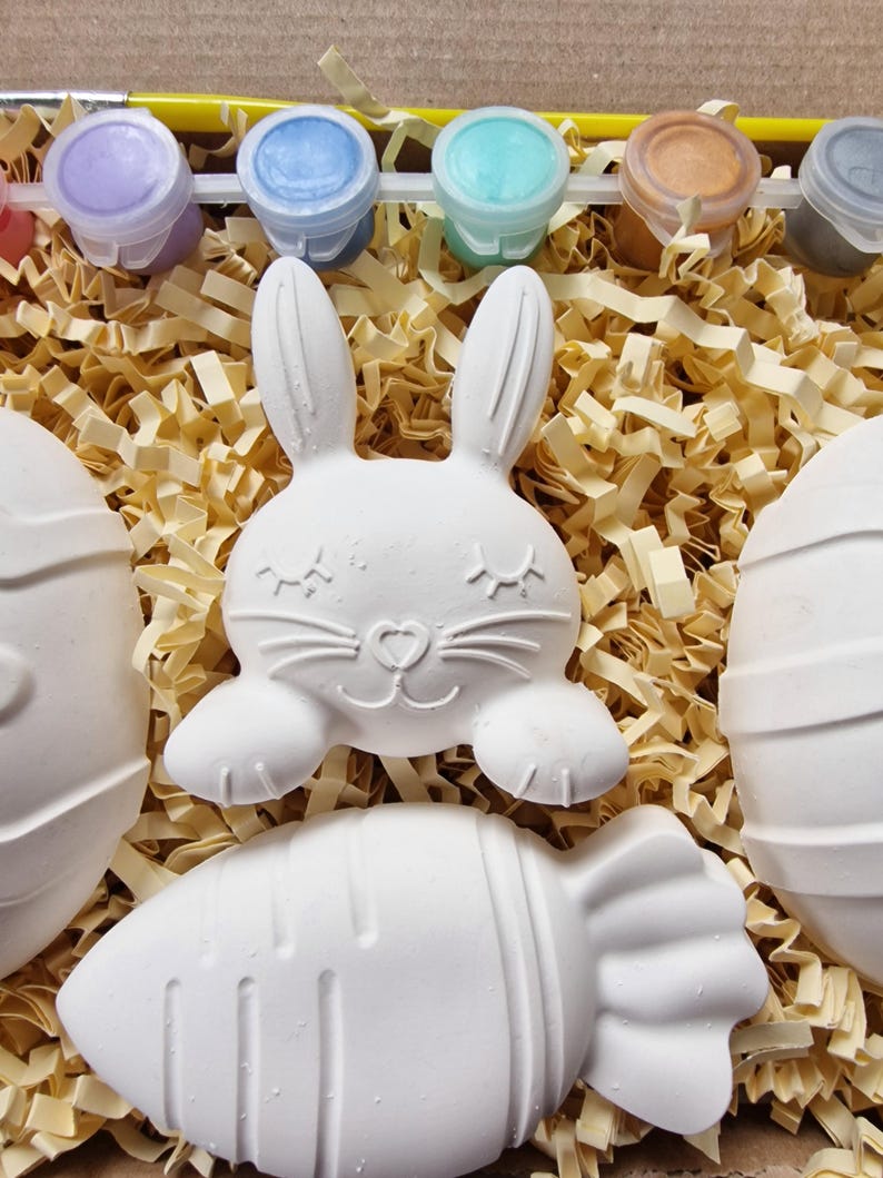 May include: A set of four white ceramic Easter-themed figurines, including a bunny, an egg, and two carrots, ready to be painted. The set comes with six small pots of paint in various colors.