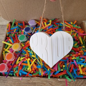 Puede incluir: Un kit de manualidades en una caja de cart&oacute;n, lleno de papel triturado de colores. El kit incluye un letrero blanco en forma de coraz&oacute;n con la palabra "PRIDE" y peque&ntilde;os botes de pintura de varios colores. Un proyecto de bricolaje creativo.