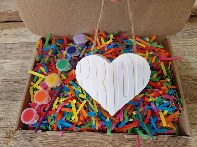 Puede incluir: Un kit de manualidades en una caja de cart&oacute;n, lleno de papel triturado de colores. El kit incluye un letrero blanco en forma de coraz&oacute;n con la palabra "PRIDE" y peque&ntilde;os botes de pintura de varios colores. Un proyecto de bricolaje creativo.