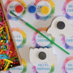 Puede incluir: Un kit de manualidades con pintura, un pincel y dos imanes de cer&aacute;mica sin pintar. El kit incluye pintura roja, azul y amarilla. Los imanes tienen forma de fantasma y cami&oacute;n. El fondo tiene el texto "KELLY'S CREATIONS".