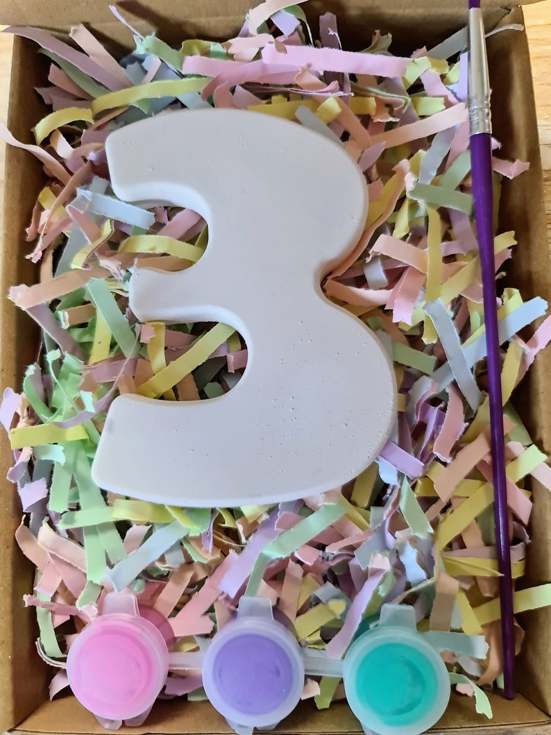 Puede incluir: Un n&uacute;mero tres de cer&aacute;mica blanca con tres peque&ntilde;os botes de pintura rosa, morado y turquesa, y un pincel morado en una caja llena de papel triturado de color pastel.