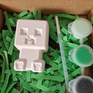 May include: A white, unpainted Creeper figure from Minecraft, ready to be painted, sits in a cardboard box filled with green paper grass. The box also contains three small paint pots with green and gray paint, and a small paintbrush.