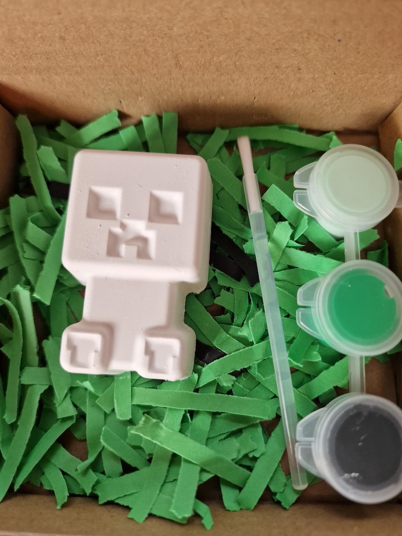 May include: A white, unpainted Creeper figure from Minecraft, ready to be painted, sits in a cardboard box filled with green paper grass. The box also contains three small paint pots with green and gray paint, and a small paintbrush.