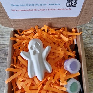 May include: A craft kit for painting a white ghost figure. The kit includes a small paintbrush, two paint pots, and the ghost figure, all nestled in orange paper strips inside a cardboard box. The box also contains instructions and a QR code.
