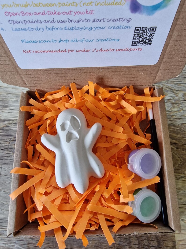 May include: A craft kit for painting a white ghost figure. The kit includes a small paintbrush, two paint pots, and the ghost figure, all nestled in orange paper strips inside a cardboard box. The box also contains instructions and a QR code.