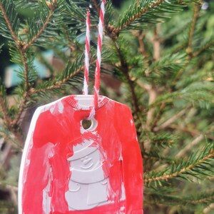 Pu&ograve; includere: Un ornamento natalizio rosso e bianco a forma di maglione con un disegno di pupazzo di neve. L'ornamento &egrave; appeso a uno spago rosso e bianco su un albero di pino verde.