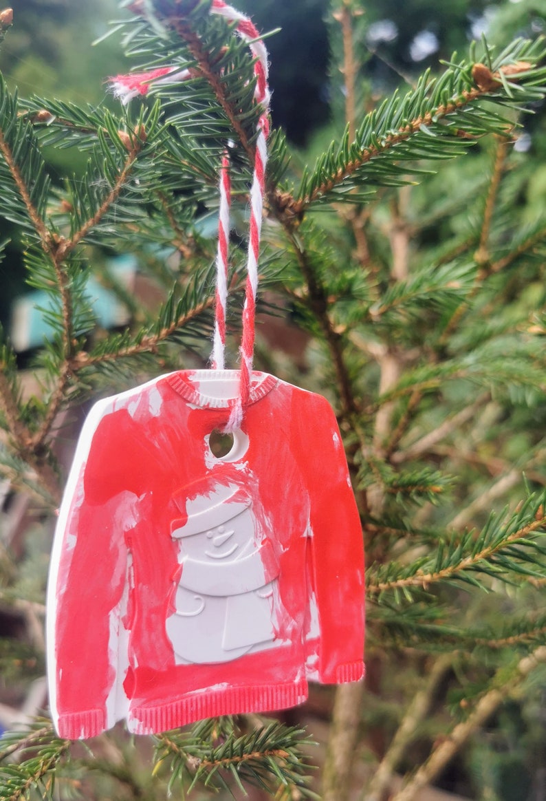 Pu&ograve; includere: Un ornamento natalizio rosso e bianco a forma di maglione con un disegno di pupazzo di neve. L'ornamento &egrave; appeso a uno spago rosso e bianco su un albero di pino verde.