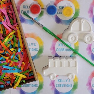 Puede incluir: Un kit de manualidades con figuras de cer&aacute;mica con forma de coche y autob&uacute;s, botes de pintura roja, azul y amarilla, y un pincel. El kit incluye una caja llena de papel triturado de colores. El fondo tiene el texto "KELLY'S CREATIONS".