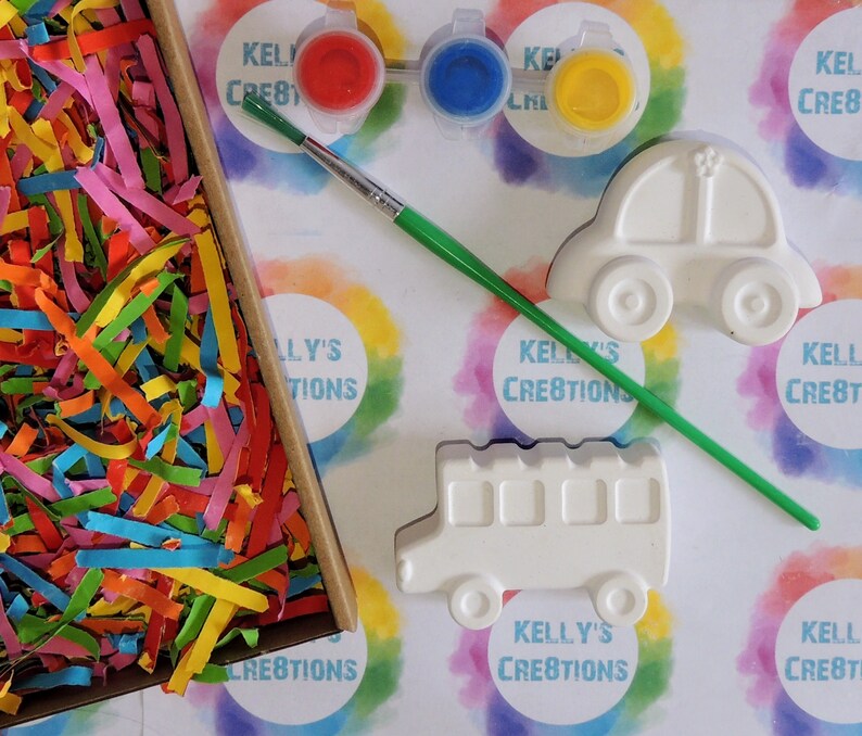 Puede incluir: Un kit de manualidades con figuras de cer&aacute;mica con forma de coche y autob&uacute;s, botes de pintura roja, azul y amarilla, y un pincel. El kit incluye una caja llena de papel triturado de colores. El fondo tiene el texto "KELLY'S CREATIONS".