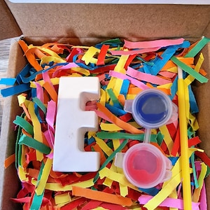 May include: A white ceramic letter E with two small pots of paint, one blue and one red, and a yellow paintbrush. The letter and paint are sitting in a box filled with colourful shredded paper. The box has a warning label that reads "Not recommended for under 3's due to small parts."