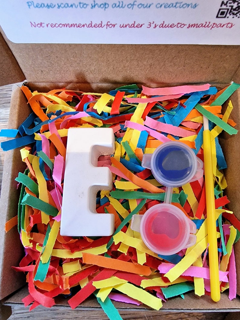May include: A white ceramic letter E with two small pots of paint, one blue and one red, and a yellow paintbrush. The letter and paint are sitting in a box filled with colourful shredded paper. The box has a warning label that reads "Not recommended for under 3's due to small parts."