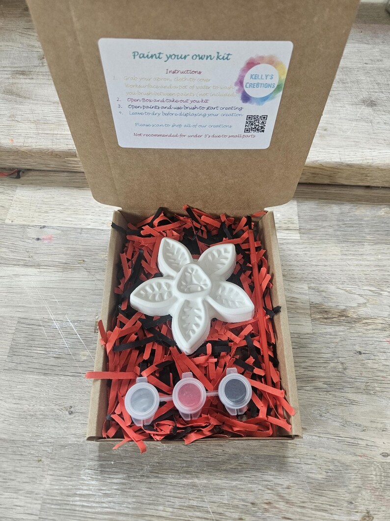 May include: A craft kit in a cardboard box containing a white flower-shaped plaster figure, three small paint pots, and red and black shredded paper filler. The box lid features a label with the text Paint your own kit and instructions.