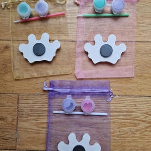 May include: Three sets of paint your own crown kits. Each kit includes a white wooden crown, two small bottles of paint, and a paintbrush. The crowns are in the shape of a traditional crown with five points. The paint colors are teal and purple, light blue and purple, and lavender and pink.