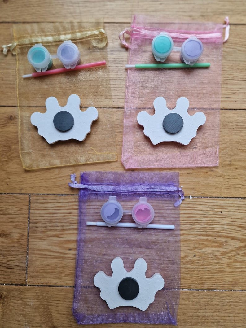 May include: Three sets of paint your own crown kits. Each kit includes a white wooden crown, two small bottles of paint, and a paintbrush. The crowns are in the shape of a traditional crown with five points. The paint colors are teal and purple, light blue and purple, and lavender and pink.