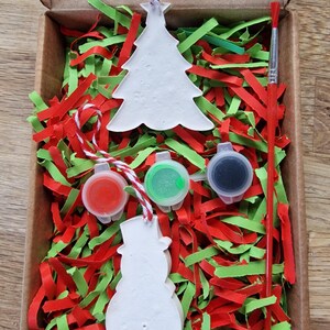 May include: A Christmas ornament craft kit with two white ceramic ornaments, one shaped like a Christmas tree and the other like a snowman. The kit includes three small paint pots, one red, one green, and one black, and a paintbrush. The ornaments are nestled in red and green shredded paper inside a cardboard box.