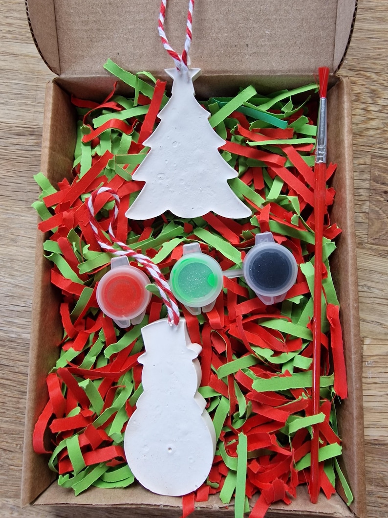May include: A Christmas ornament craft kit with two white ceramic ornaments, one shaped like a Christmas tree and the other like a snowman. The kit includes three small paint pots, one red, one green, and one black, and a paintbrush. The ornaments are nestled in red and green shredded paper inside a cardboard box.