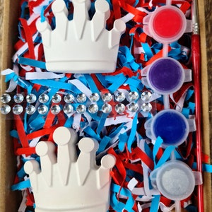 May include: A craft kit containing two white ceramic crowns, paint pots in pink, red, purple, blue, silver, and yellow, and a paintbrush. The crowns and paint pots are in a box with red, white, and blue shredded paper.