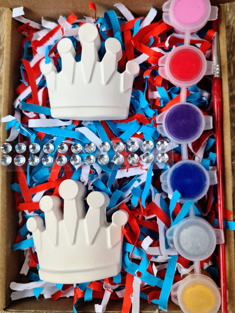 May include: A craft kit containing two white ceramic crowns, paint pots in pink, red, purple, blue, silver, and yellow, and a paintbrush. The crowns and paint pots are in a box with red, white, and blue shredded paper.