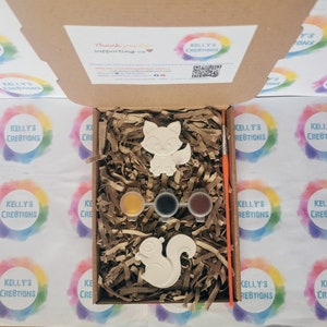 May include: A craft kit with two ceramic animal figurines, a fox and a squirrel, ready to be painted. The kit includes three small paint pots, a paintbrush, and brown packing peanuts.