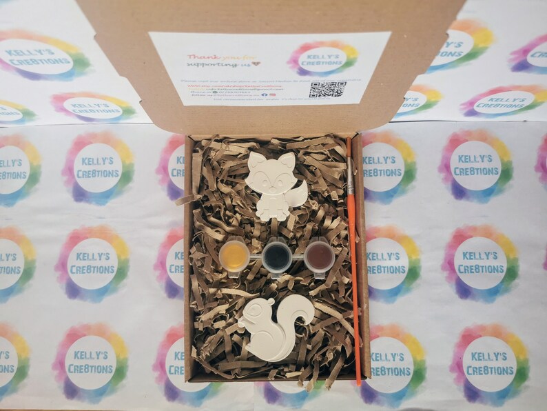 May include: A craft kit with two ceramic animal figurines, a fox and a squirrel, ready to be painted. The kit includes three small paint pots, a paintbrush, and brown packing peanuts.