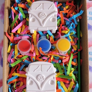 May include: A craft kit with two white ceramic Volkswagen bus figurines, three paint pots, and a paintbrush. The paint pots are red, blue, and yellow. The kit is packaged in a cardboard box with colorful shredded paper.