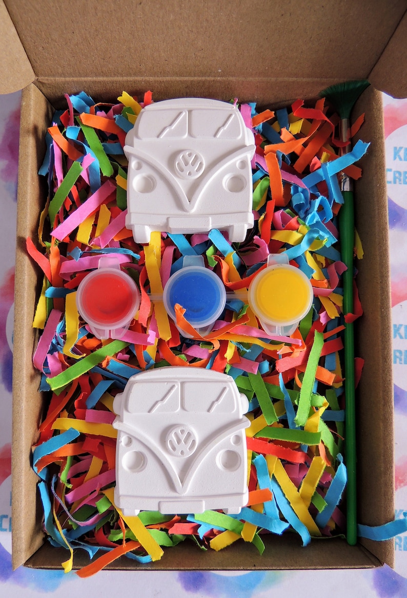 May include: A craft kit with two white ceramic Volkswagen bus figurines, three paint pots, and a paintbrush. The paint pots are red, blue, and yellow. The kit is packaged in a cardboard box with colorful shredded paper.