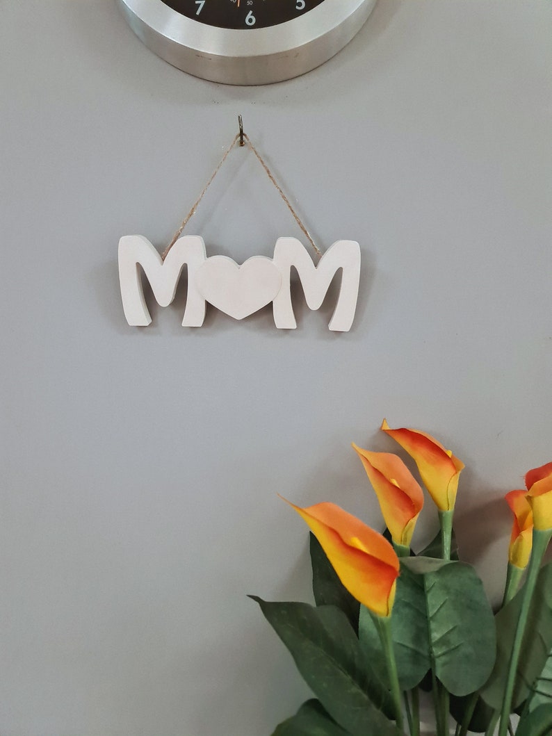 Pu&ograve; includere: Un cartello di legno bianco con la parola "MOM" scritta con un cuore al posto della "O". Il cartello &egrave; appeso a una corda ed &egrave; contro un muro grigio. Ci sono fiori arancioni nell'angolo in basso a destra dell'immagine.
