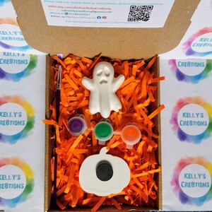 May include: A Halloween-themed craft kit in a cardboard box. The kit includes a white ghost and pumpkin, paint pots in purple, green, and orange, and a paintbrush. The box is filled with orange paper shreds.