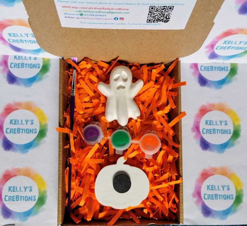 May include: A Halloween-themed craft kit in a cardboard box. The kit includes a white ghost and pumpkin, paint pots in purple, green, and orange, and a paintbrush. The box is filled with orange paper shreds.