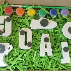 May include: A white wooden craft kit with the word "GOAL" spelled out in large letters. The kit includes a trophy, a soccer shoe, and a circle. The kit also includes six small paint pots with red, orange, yellow, blue, purple, and green paint.