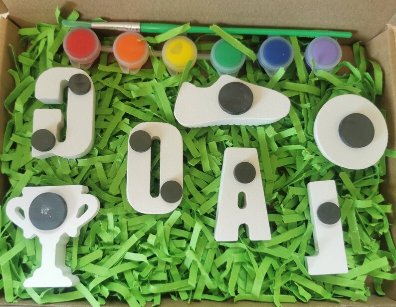 May include: A white wooden craft kit with the word "GOAL" spelled out in large letters. The kit includes a trophy, a soccer shoe, and a circle. The kit also includes six small paint pots with red, orange, yellow, blue, purple, and green paint.