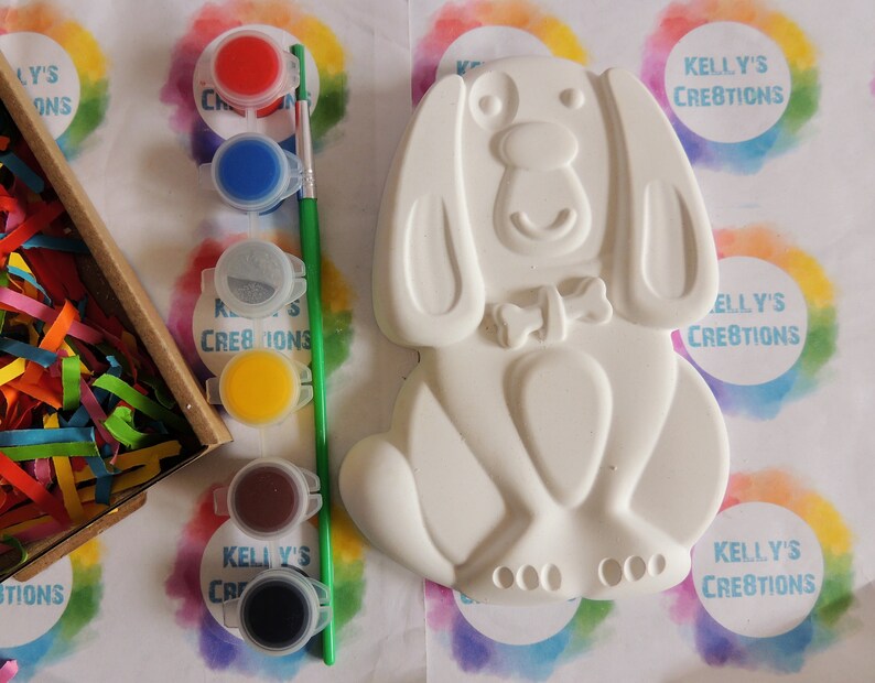 May include: White ceramic dog figurine with a bone on its collar. The figurine is ready to be painted with six small pots of paint in red, blue, gray, yellow, brown, and black.