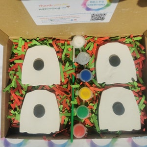 May include: A box filled with green and red shredded paper containing four white ceramic ghost shapes with black holes for eyes and a set of five paint pots in white, gray, blue, yellow, and red.  The box lid is open and has a thank you message and a QR code.