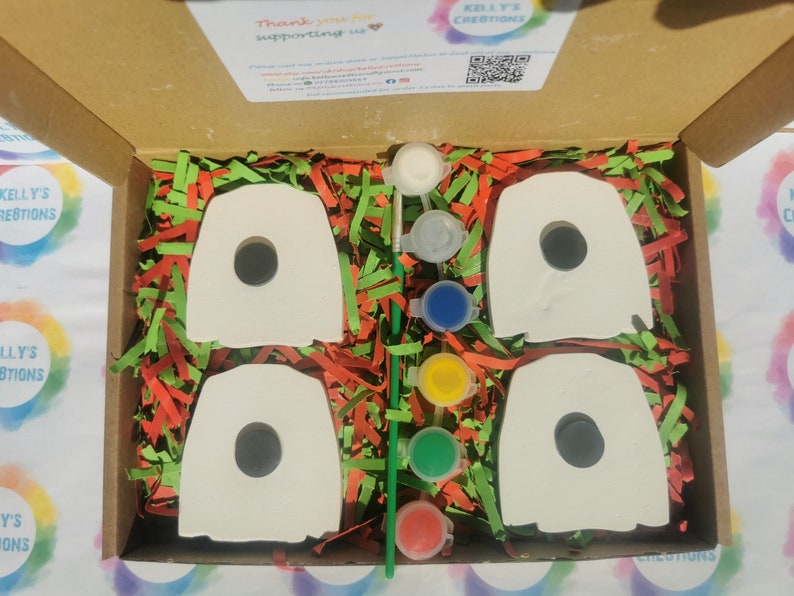 May include: A box filled with green and red shredded paper containing four white ceramic ghost shapes with black holes for eyes and a set of five paint pots in white, gray, blue, yellow, and red.  The box lid is open and has a thank you message and a QR code.