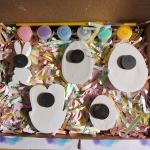 May include: A craft kit for painting Easter decorations. The kit includes five white ceramic pieces shaped like eggs and a bunny, five small paint pots in pink, purple, blue, green, and yellow, and a paintbrush. The pieces are nestled in shredded paper inside a cardboard box.