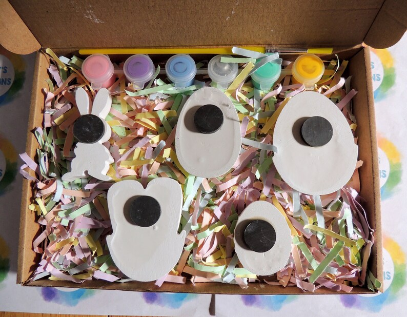May include: A craft kit for painting Easter decorations. The kit includes five white ceramic pieces shaped like eggs and a bunny, five small paint pots in pink, purple, blue, green, and yellow, and a paintbrush. The pieces are nestled in shredded paper inside a cardboard box.