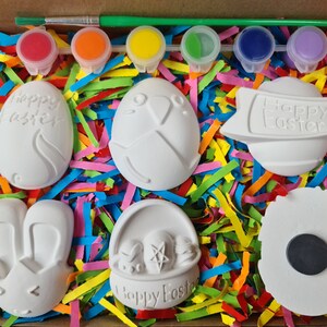 May include: A set of six white ceramic Easter-themed ornaments in a box with colourful shredded paper. The ornaments include a bunny, an egg with the words "Happy Easter", a basket, a chick, a banner with the words "Happy Easter", and a plain egg. The set also includes five small paint pots and a paintbrush.