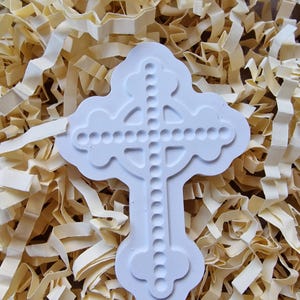 May include: White ceramic cross mold with a beaded design. The mold is sitting on a bed of shredded paper.