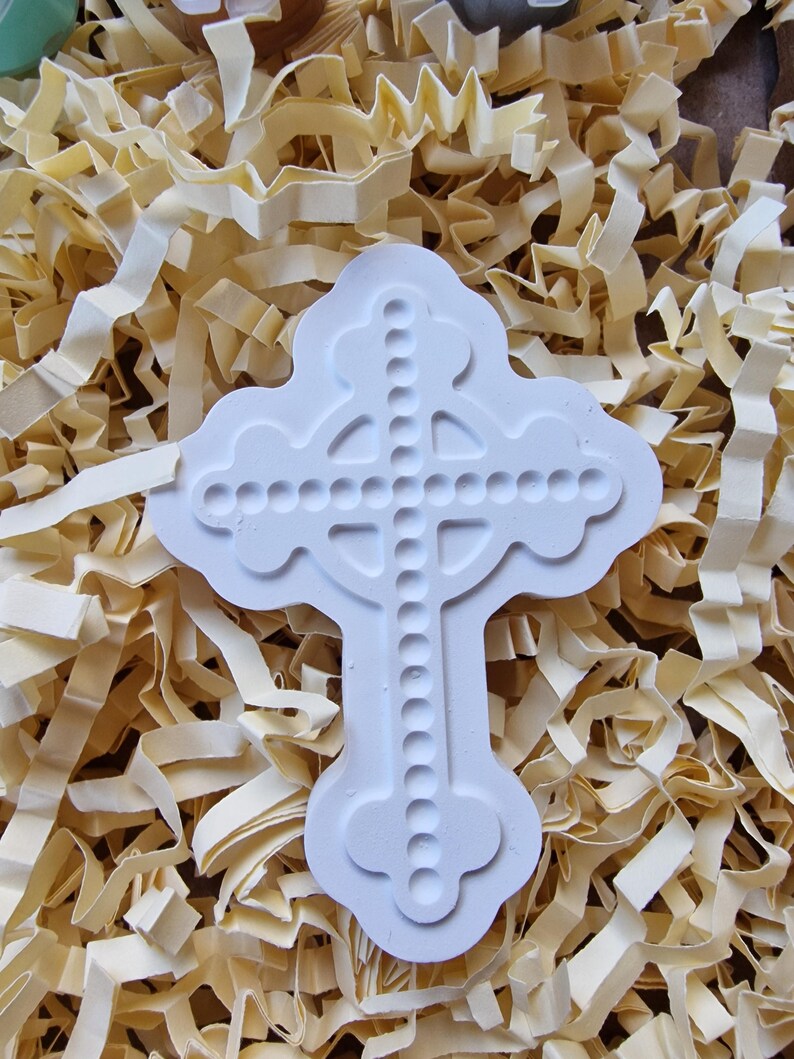 May include: White ceramic cross mold with a beaded design. The mold is sitting on a bed of shredded paper.