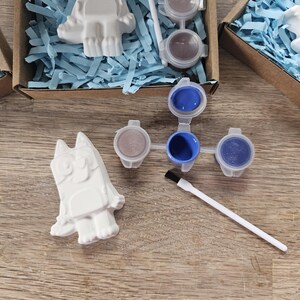 May include: A craft kit with cartoon character figurines, paint pots, and paintbrushes. The white figurines sit in cardboard boxes with blue paper. The paint pots contain various colours and are arranged on a wooden surface. The kit is for painting.