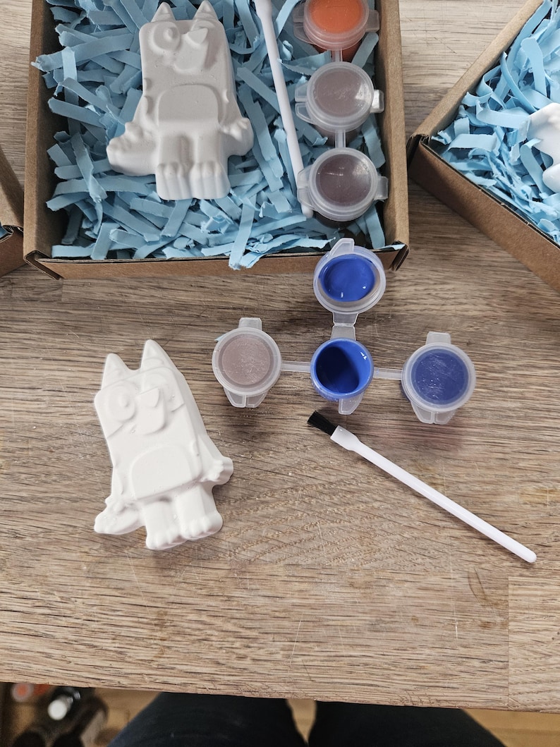 May include: A craft kit with cartoon character figurines, paint pots, and paintbrushes. The white figurines sit in cardboard boxes with blue paper. The paint pots contain various colours and are arranged on a wooden surface. The kit is for painting.