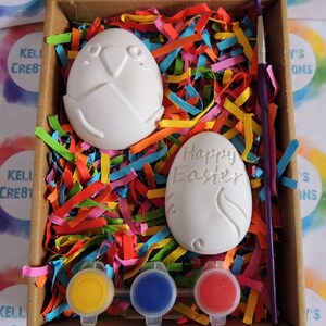 May include: A craft kit for painting ceramic Easter eggs. The kit includes two white ceramic eggs, one with the words "Happy Easter" and the other with a bird design, three small pots of paint in yellow, blue, and red, and a paintbrush. The eggs are nestled in colourful shredded paper inside a cardboard box.