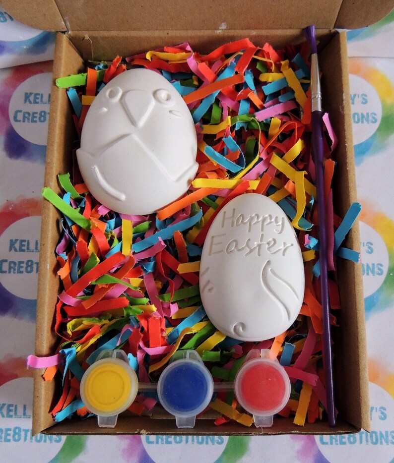 May include: A craft kit for painting ceramic Easter eggs. The kit includes two white ceramic eggs, one with the words "Happy Easter" and the other with a bird design, three small pots of paint in yellow, blue, and red, and a paintbrush. The eggs are nestled in colourful shredded paper inside a cardboard box.