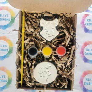 May include: A craft kit in a cardboard box, featuring a Hufflepuff crest and a Harry Potter logo. The kit includes three paint pots in black, yellow, and red, a yellow paintbrush, and two blank circular and shield-shaped objects. The box is filled with brown paper shreds.