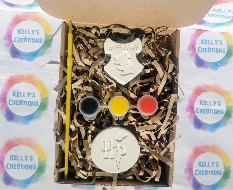 May include: A craft kit in a cardboard box, featuring a Hufflepuff crest and a Harry Potter logo. The kit includes three paint pots in black, yellow, and red, a yellow paintbrush, and two blank circular and shield-shaped objects. The box is filled with brown paper shreds.